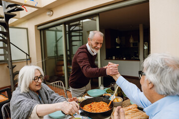 Two male friends shake hands over a table with a female friend sitting behind them while they smile