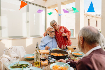 A woman stands and holds a glass while hugging a man who is sitting at a table next to a man and a...
