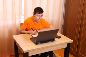 Schoolboy taking notes while working with laptop in room.