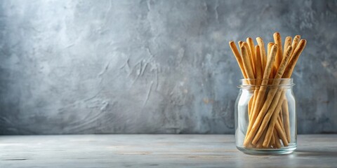 A glass jar filled with crunchy breadsticks rests on a rustic surface against a textured backdrop