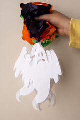 Vertical shot of hand holding colorful paper craft, including ghost cutouts, with vibrant floral designs against a plain wall.