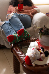 Unrecognizable woman lounging on a sofa, wearing festive Christmas socks, browsing her phone, surrounded by a warm and cozy atmosphere