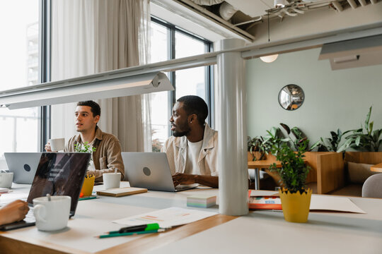 Two male workers sit at desks in front of laptops while one of them smiles and the other holds a cup