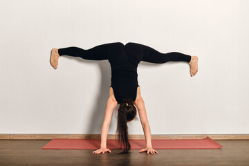 Woman performs inverted yoga pose against white wall in tranquil studio. Red mat contrasts with wooden floor, creating balance. Natural light enhances calm ambiance