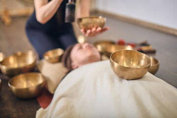 Young woman receives sound healing therapy with singing bowls. Warm lighting enhances relaxation...