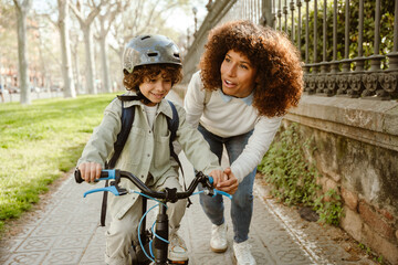 A mother walks and holds the bicycle on which her smiling son rides
