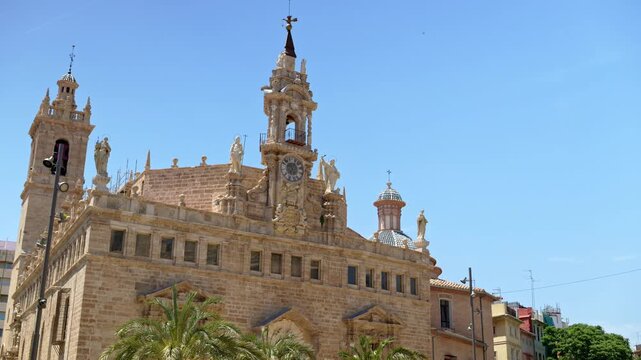 Low angle view of the baroque church Sant Joan del Mercat with statues, central clock and belfry, and blue-tiled domes against a bright blue sky in Valencia, Spain