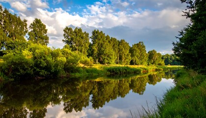 Fototapeta premium Calm river reflects trees under a partly cloudy sky. Gentle light shines