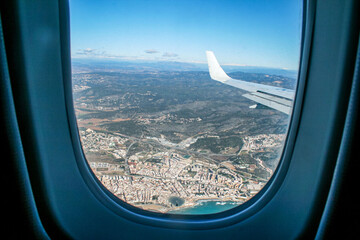 Naklejka premium Aerial view from airplane window: city, coast and mountain landscape under clear blue sky. Travel, vacation, tourism, transportation and adventure concept.