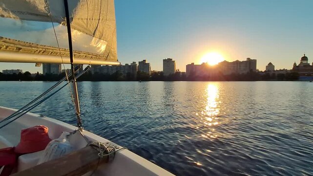 Yacht with a sail sailing along the river. Beautiful sunset. Evening yacht ride against the backdrop of the setting sun. Sun sets behind the houses on the horizon. Yacht is rocking on the waves