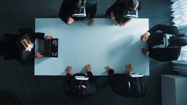 Top down aerial view of diverse data analysts wearing VR headset and stand around table while using hand gestures to manipulate the data streamed live from executive manager's laptop. Directorate.