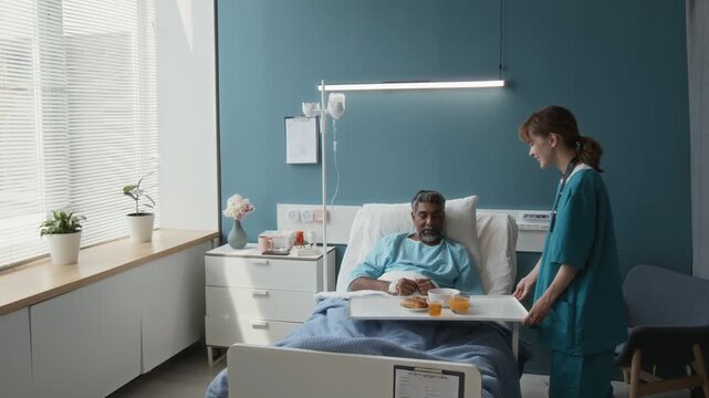 Positive young nurse bringing meal for middle-aged ill man sitting in bed with IV drip while undergoing treatment in hospital ward