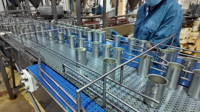 Industrial Canning Line: A detailed view of a food processing plant, showcasing a conveyor belt system in motion with numerous tin cans moving along, under the supervision of a worker.