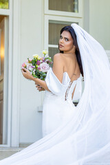 Bride in elegant lace wedding dress holding floral bouquet standing gracefully in front of historic mansion courtyard captured in golden hour light for fine art bridal photography
