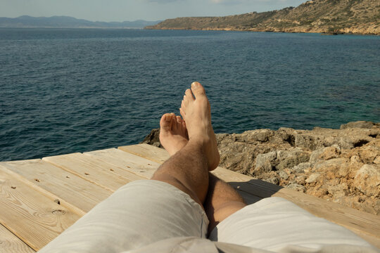 View of a man's feet resting on a coastal wooden path by the Mediterranean Sea on a sunny summer day, concept of vacation, relaxation and lifestyle