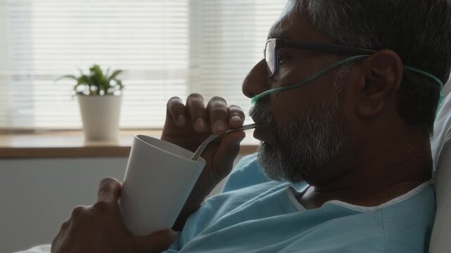 Side view of mature man wearing glasses and hospital gown sipping drink through straw while receiving oxygen through nasal cannula