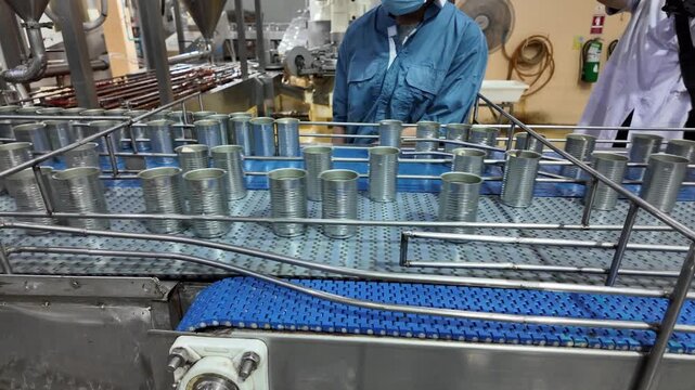Automated Food Production Line: Cans move along a conveyor belt in a sterile food processing factory, with workers in protective gear overseeing the automated assembly process.