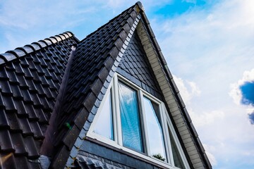 Modern triangular roof with black tiles and white window reflecting blue sky © Michael