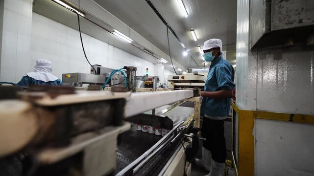Food Production Facility: A worker wearing a mask and hat operates machinery in a brightly lit food processing plant, focusing on efficient production.