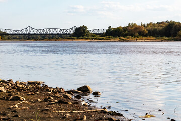 Riverbank with rocks and distant truss bridge under blue sky