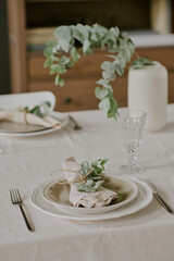 Elegant table setting featuring plate with linen napkin tied with twine and eucalyptus branch, glass goblet and cutlery arranged on tablecloth, ceramic vase with greenery in background
