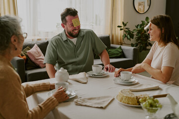 Caucasian middle aged man sitting at table with sticky note on forehead laughing with woman and senior woman drinking tea and talking together