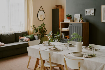 Modern dining room featuring set table with plates, glasses, cutlery and decorative plants on tablecloth, background showing sofa, shelving unit, framed art and large window with sheer curtains