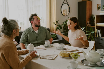 Caucasian senior woman, Caucasian middle aged man, Caucasian middle aged woman sitting at dining table playing guessing game with sticky notes on foreheads, smiling and gesturing