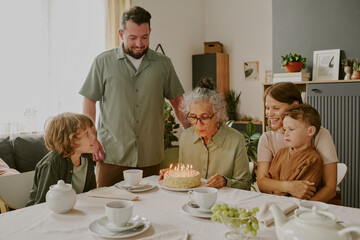 Senior Caucasian woman blowing out candles on birthday cake surrounded by her family at dining table