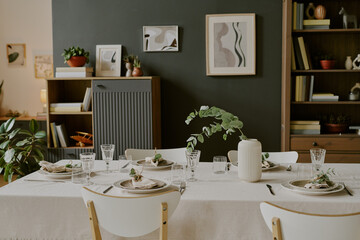Modern dining room table set for meal with glassware, plates, cloth napkins, and ceramic vase with greenery, contemporary wall art and plants decorating background shelves