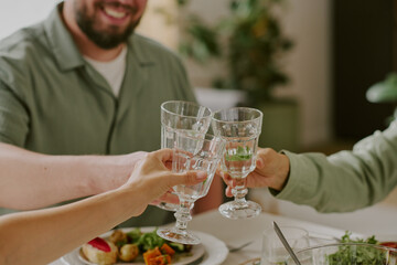 Three young adults clinking glasses while sitting at table, celebrating together, hands and partial faces visible, sharing meal