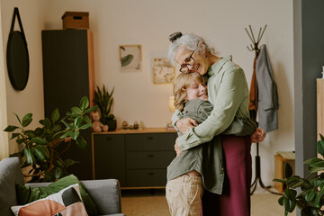 Senior Caucasian woman embracing smiling Caucasian child in cozy living room, both standing and showing affection, plants and wall decor visible in background