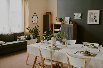 Modern dining room featuring table set for meal with six plates, glasses, cutlery, and decorative plants, cozy living area with sofa and indoor plants visible in background