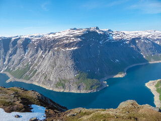 An immense, snow-capped mountain massif with rugged granite cliffs rises majestically above the deep blue waters of the Ringedalsvatnet fjord on a clear day near Trolltunga, Norway's famous hike.
