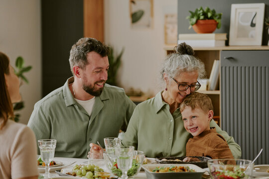 Caucasian senior woman embracing smiling child while sitting at dining table with young Caucasian man, family members gathering and sharing meal together indoors