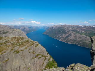 A stunning panoramic vista shows the deep blue waters of the Lysefjord winding between immense, rocky mountains, viewed from the iconic Preikestolen hiking area in Norway under a clear summer sky.