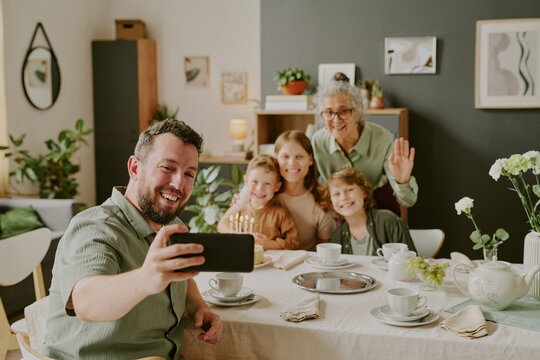 Caucasian young adult man taking selfie with smiling Caucasian middle aged woman, senior woman, and two Caucasian children sitting at dining table celebrating birthday together - Powered by Adobe