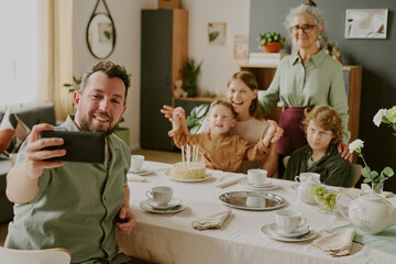 Caucasian young adult man taking selfie with family celebrating birthday at dining table