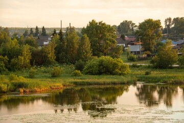 Fototapeta premium Beautiful lake in summer. Lush greenery, houses in the background, clouds, reflection.