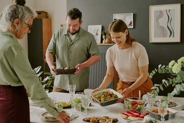 Middle aged Caucasian woman serving food at dining table with middle aged man holding bread basket and senior woman arranging tableware during family meal