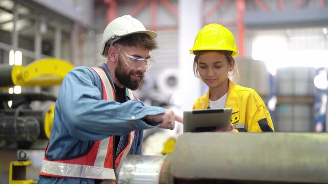 Factory Collaboration: A seasoned male factory worker, wearing a hard hat and safety glasses, guides a young female apprentice using a tablet.