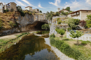 Fototapeta premium Natural rock arch over a serene river - A river flows through a natural rock arch, with ancient houses perched on the cliff above under a blue sky.