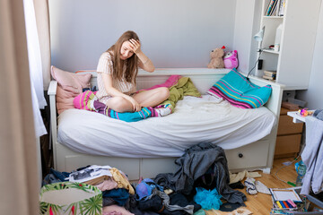 A teenage girl in pajamas sits in bed, looking shocked at the mess in her cluttered bedroom