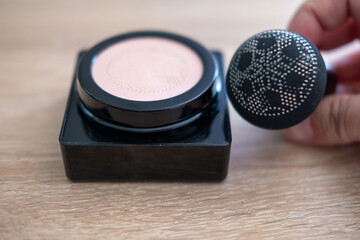 black makeup sponge with dotted pattern, poised above open compact powder case on wooden surface, highlighting beauty and skincare tools, daily skincare and grooming, Cosmetic product, daily use