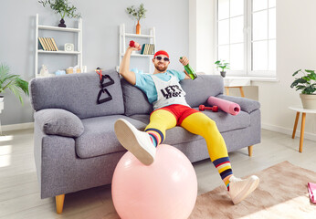 Funny man in sportswear sits on the couch, drinking alcohol during home gym training. This lazy twist on a workout adds a comical touch to home sport routine, blending relaxation and exercise.