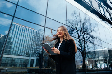 Confident businesswoman smiling while holding tablet outdoors near modern glass building reflecting trees and sky, professional lifestyle concept