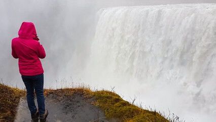 A female tourist in a bright pink raincoat stands at the precipice, photographing the immense and powerful Dettifoss waterfall as it disappears into a thick cloud of mist in Iceland's wilderness.
