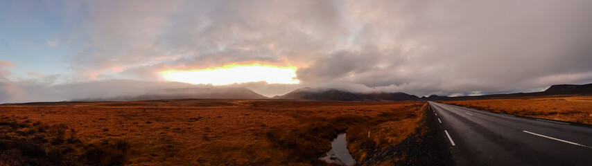 A dramatic panoramic view of an empty road running through a vast, golden tundra in Iceland, as a powerful sunset breaks through heavy clouds, casting a warm, epic glow over the misty mountains.