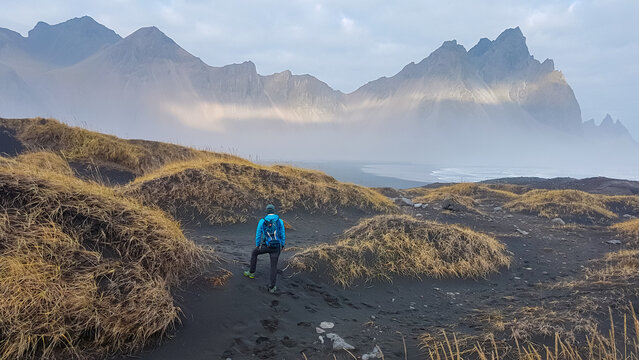A female hiker with a backpack walks through black sand dunes towards the coast, as a dramatic sunbeam illuminates the majestic, misty Vestrahorn mountain in the background at Stokksnes, Iceland. - Powered by Adobe