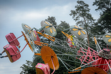 Bright pink and yellow carnival ride swings high in the air, with flower-shaped tops spinning against a blue sky and trees in the background.